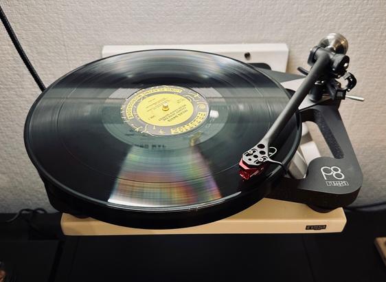 Photo of a record spinning on the skeletal platter of a Rega P8 turntable. 

The centre label is yellow with just the words, Miles Davis, legible above the spindle. 

The tone arm and red cartridge are tracking the opening track.