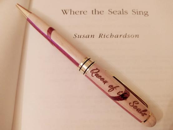 A wooden pen, with the words 'Queen of the Seals' etched along it, rests on a copy of 'Where the Seals Sing' by Susan Richardson.