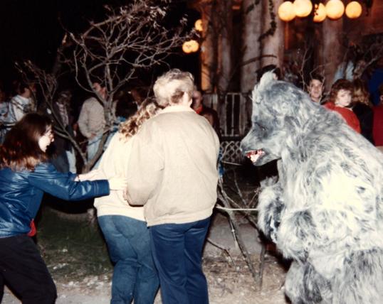 Large grey werewolf (Snarl, worn by Ysengrin) interacting (chasing) two guests in a queue line for a haunted house (haunt facade is partially visible in background). The two guests are holding each other as they run, and are trying to shield themselves behind a third guest. Other patrons watch in the background.