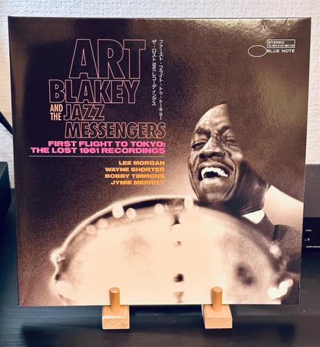 Photo of a record album cover displayed on a wooden stand. 

The cover is a photo of Art Blackley playing the drums. The photo is a close up, we can see his face - grinning in concentration - framed between the crash cymbal and tom tom. 

The background is smoky black and, in the top left hand corner, printed in yellow, ‘Art Blackey and the Jazz Messengers’. Then, below this, in pink typeface, ‘First Flight to Tokyo: The Lost 1961 Recordings’.

Along the righthand edge of this text Japanese lettering runs vertically down. 

Beneath this are listed, in orange type, ‘Lee Morgan, Wayne Shorter, Bobby Timmons, Jymie Merritt’.