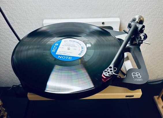 Photo of a record spinning on the skeletal plinth of a Rega P8 turntable. 

The centre label, blue and white, reads, ‘Blue Note’. The remainder of the lettering is lost in motion blur. 

The tone arm is extended and tracking the first song.