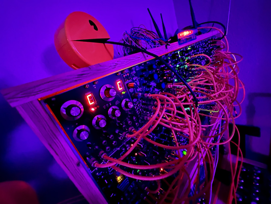 wide angle photo of a eurorack case made of splintered plywood. it is populated by black panel modules with yellow cables. the cables look red in the lighting. Atop it sits a pacman phone commanding attention by chewing on a midi cable. behind pacman is a nest of cables and a wifi pineapple. the lighting is a dramatic blue and purple.