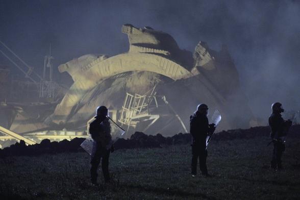 A spaced out row of riot police standing on grass, silhouetted against an enormous mining machine in the background.