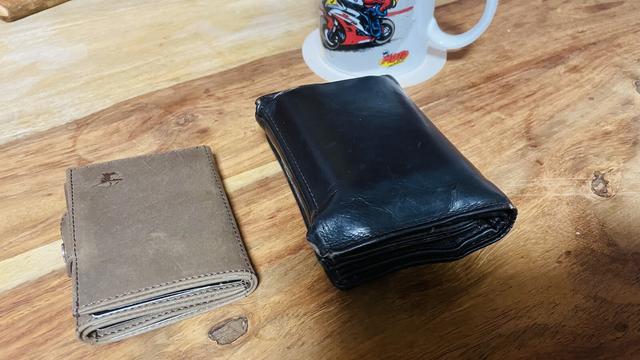Man sieht auf dem Bild zwei Geldbörsen auf einem Holztisch liegen. Links eine neue, hellbraune Geldbörse, rechts die alte, schwarze die deutlich größer ist.