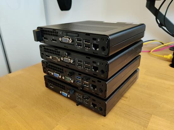 Four HP Prodesk Minis machines stacked on top of each other on a desk, back view.