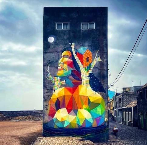 Street Art Wall. A four-story rectangular tower-like building was sprayed with a colorful mural. The background is black and makes the figure in the foreground shine. In cubist Popartstyle, a figure with a cap, painted from colorful rectangles and triangles, can be seen. She looks to the left into the sky. From her right shoulder grows a striped tree with a brick tree house on it. A large black bird, with stars for plumage, sits in front of it. Another colorful tree can be seen to the left side. Above them all is a white moon.