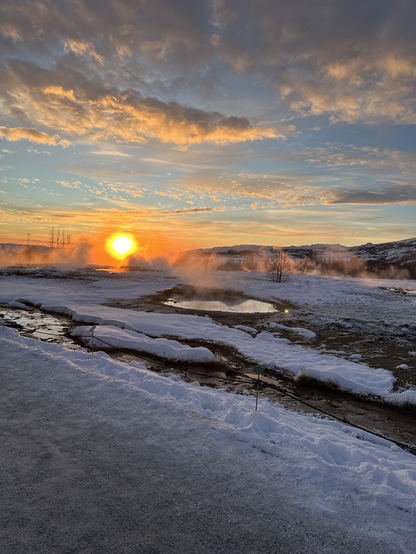 Sunset over snowy hills, with hot steam rising over small pools of water.