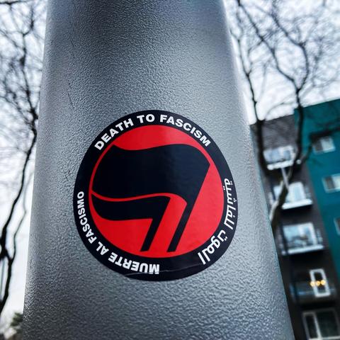 Red and black round sticker with two black “antifascist” flags in the middle surrounded by the words “death to fascism” in three languages, posted on a gray utility pole outdoors with trees in the background