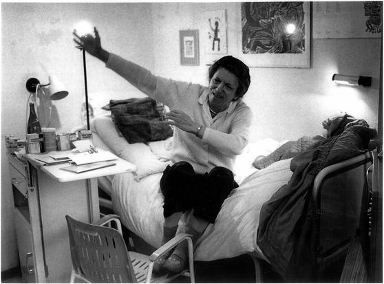 Historische Fotoaufnahme der Dichterin Rose Ausländer in späteren Jahren. Sie sitzt auf einem Bett und gestikuliert mit den Armen. Ihre Füße hat sie auf einem Stuhl abgelegt. Während das Foto aufgenommen wurde, war die Dichterin im Gespräch mit dem ehemaligen Stern-Journalisten Jürgen Serke. Dieser ist auf dem Bild nicht zu sehen.