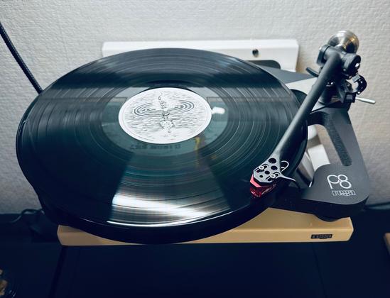 Photo of a record spinning on the skeletal plinth of a Rega P8 turntable.

The centre label is patterned with a black sketched drawing, the subject unidentifiable. 

The tone arm, red cartridge, traces the first track.