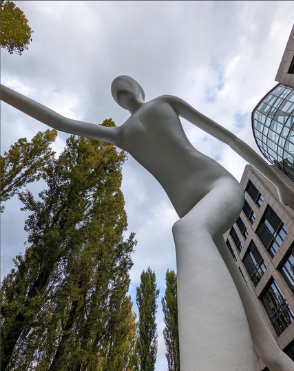 Au pied d'une statue d'un homme en train de marcher, le bras droit en avant, le bras gauche en arrière, la jambe gauche en avant, la jambe droite en arrière. Cet homme est complètement blanc et ne possède pas de visage.