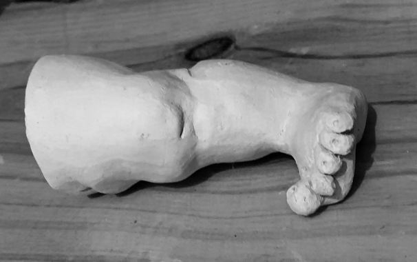 black and white photograph of a doll's leg modeled from clay.  it is planned as a part for a mobile with doll body parts and therefore has a small hole at the top edge for hanging.  the leg is fleshy and thick and has a short, wide foot with rounded, splayed toes.  picture 1: front view with knee.