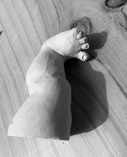 black and white photograph of a doll's leg modeled from clay.  it is planned as a part for a mobile with doll body parts and therefore has a small hole at the top edge for hanging.  the leg is fleshy and thick and has a short, wide foot with rounded, splayed toes.  picture 2: side view of the outside
