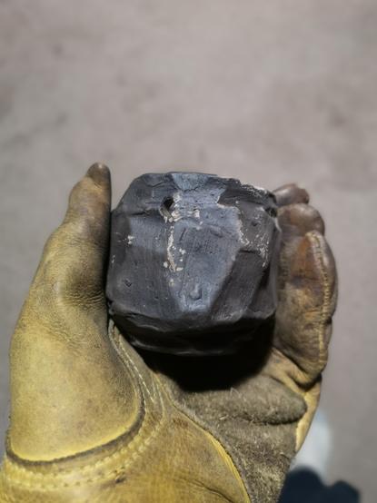 A small roughly cut ceramic container held in a leather glove. The item is a dark smokey colour and has some ash on it.