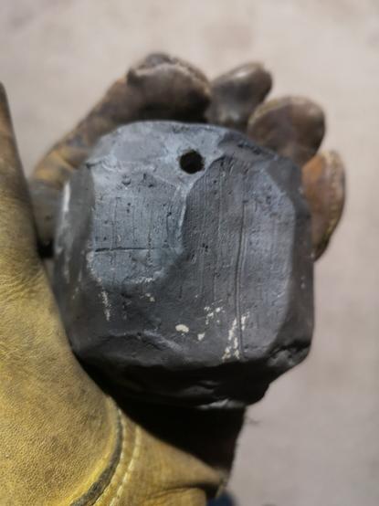 A small pitfired stoneware pot held in a leather glove. The surface is smokey grey and has visible cut marks.