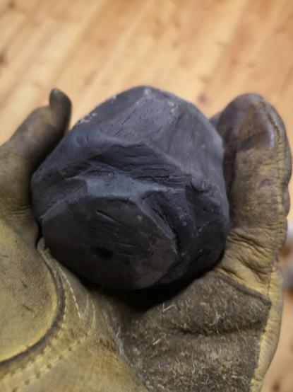 A small pitfired stoneware clay pot held in a leather glove, viewed from underneath. There are visible rough carving marks in the surface, which is a dark smokey colour from the pit fire.