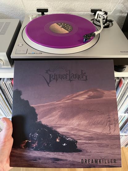A purple vinyl record spinning on a silver technics player. Below there’s the cover of Sumerlands Dreamkiller. It has the band logo on top and there’s a burning car wreck in the desert with dunes in the back.