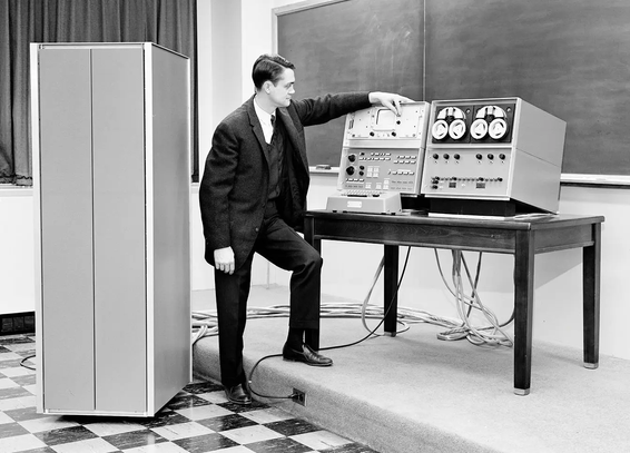 A black and white photo of a classroom, looking diagonally to the corner between the windows (curtains drawn) and the empty chalkboard.  There is a short step up that forms a stage in front of the chalkboard, from which comes an electrical power lead to the two modules (one for front panel switches and oscilloscope display, and one for the LINCTape drives) that sit on a table on stage in front of the chalkboard, along with the keyboard.  Bulky data cables run from these modules along the stage floor to a cabinet the size of a refrigerator that sits down on the chequered floor in front of the stage.
A slender white man with dark hair combed in a slick part and a well-tailored dark three-piece suit stands with one foot on the chequered floor and one up on the stage.  His left arm is draped leisurely over the oscilloscope console, his fingers reaching somewhat awkwardly  to pinch a contrast dial on the scope.  His right arm dangles at his side rather than reach for the keyboard, and he gazes longingly into the scope's display.