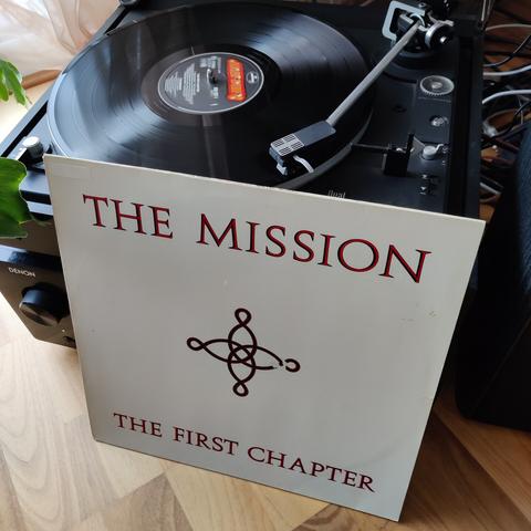 Schallplatte von The Mission - The First Chapter wird auf einem schwarzen Dual-Plattenspieler abgespielt. Davor steht die Plattenhülle mit Cover in Weiß und roter Schrift, oben der Bandname, unten der Albumtitel in Versalien mit Serifen. Mittig ein senkrechtes, gleichschenkeliges Kreuz aus verschnörkelten roten Ornamentlinien.
