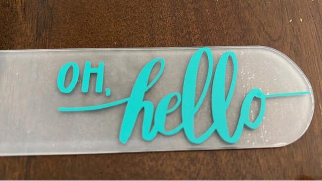 Phrase “oh hello” is a vinyl sticker placed on an acrylic bookmark. Rounded end.