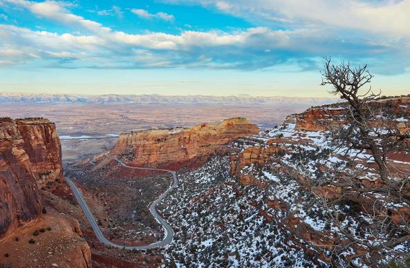 Colorado National Monument landscape photo with cliffs in the foreground, a winding road at the bottom of a canyon, and mountains in the distance.