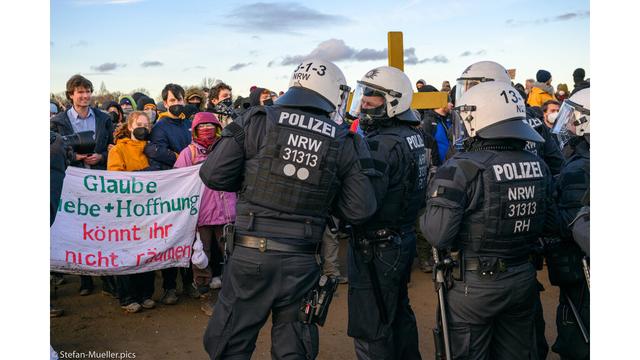 Aktivisten mit einem gelben Holzkruez und einem Banner „Glaube, Liebe + Hoffnung könnt Ihhr nicht räumen“ stellen sich Polizei entgegen. Lützerath, 08.01.23