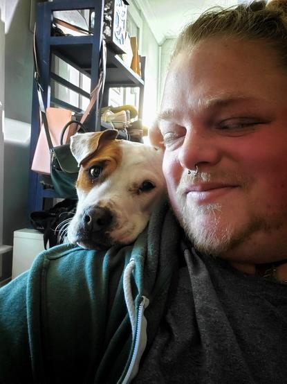 A goofy white & brown pit bull mix rests her shoulder on an equally goofy nonbinary dork. The enby gazes with affection and frustration at the dog.