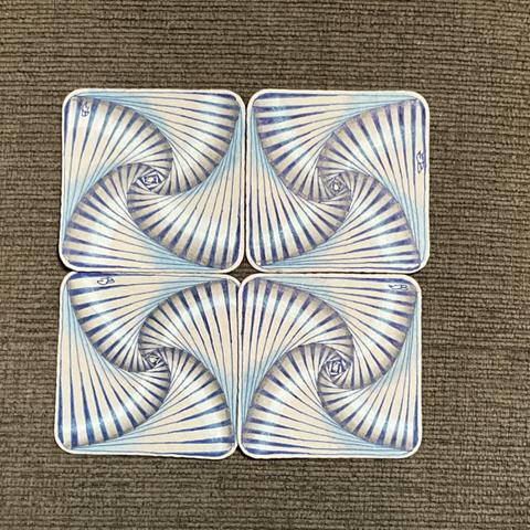 4 grey Zentangle Bijou tiles are placed in a square on a dark grey surface. On each tile is the same geometric line pattern in dark blue, which looks like it is spiralling into the centre of the tile. Graphite shading is added to make the spiral look more 3D, and the colours are boosted with white and light blue chalk pastel. These 4 tiles are positioned and look like fans are coming together across the tiles
