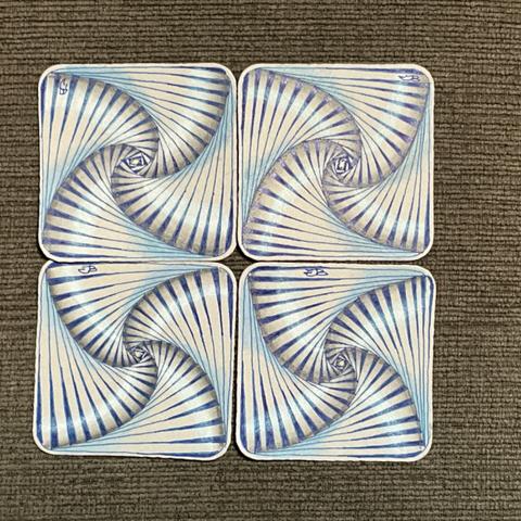 4 grey Zentangle Bijou tiles are placed in a square on a dark grey surface. On each tile is the same geometric line pattern in dark blue, which looks like it is spiralling into the centre of the tile. Graphite shading is added to make the spiral look more 3D, and the colours are boosted with white and light blue chalk pastel. These 4 tiles are positioned so that it looks like they are spiralling into each other in tubes and fans shapes