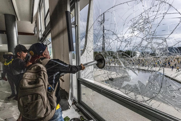 A photograph of a supporter of Jair Bolsonaro smashing a window within a government building in Brasília on January 8, 2023.