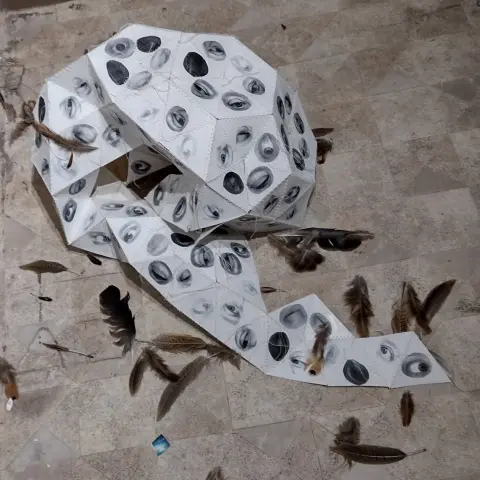Collage sculpture of a collapsed geodesic sphere on brown gallery floor. The sphere consists of numerous triangles depicting tired looking eyes, one in each triangle. The black ans white structure is surrounded with scattered feathers.