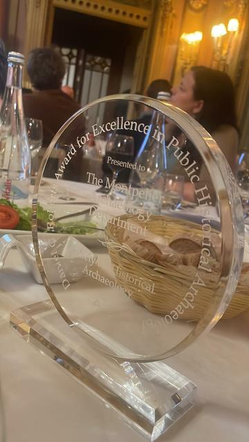 A clear, circular award with a rectangular base sits on a banquet table. The award reads, " Daniel G. Roberts Award for Excellence in Public Historical Archaeology/ Presented to The Montpelier Archaeology Department/ Society of Historical Archaeology"