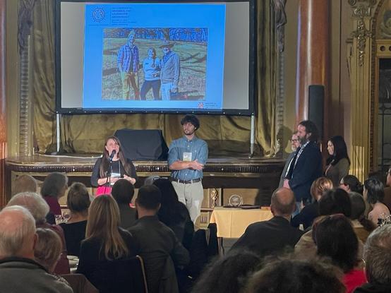 Two people stand center stand, a woman on the left, speaking into a microphone and a man to her left, standing with arms crossed. To his left there are three other people standing, looking towards them. A full audience is before them. Behind the 2 speakers is a projected image of 3 people in a field smiling.