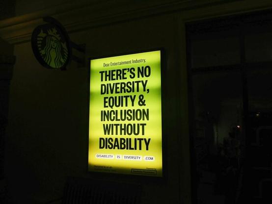 Image of a Yellow and Green lit cinema style sign on the side of a building hanging below another sign of a well known shop selling overpriced coffee (I think you can guess which coffeshop).  Black lettering on the cinema sign reads:

"Dear entertainment industry. THERE'S NO DIVERSITY, EQUITY & INCLUSION WITHOUT DISABILITY.  DISABILITY IS DIVERSITY .COM"