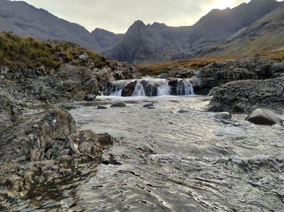 A calm pool beneath a shallow waterfall with a mountainous backdrop