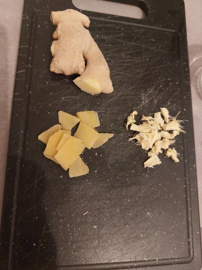 A black chopping board with a ginger root alongside some pale yellow slices of cooked ginger and light colored dried ginger