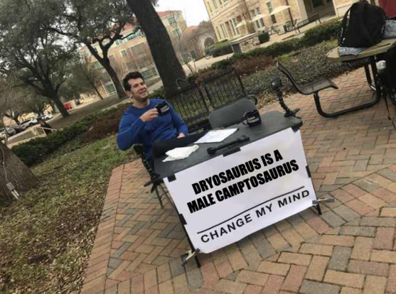 A smug man sits behind a desk. The sign on the front says "Dryosaurus is a male Camptosaurus – change my mind".