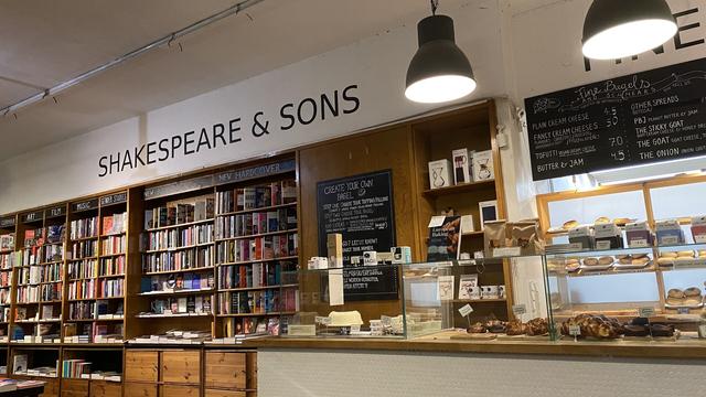 On the left side, a wall of bookshelves; on the other, a cafe counter. A sign on the wall above them say: Shakespeare & Sons