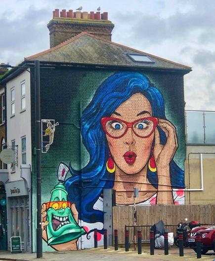 Street art wall.
Mural on the outer wall of a two-story commercial building. In bright colors is sprayed portrait of a woman in comic style. The background is green. She has blue hair and put her right hand to her red glasses as if she wants to see something better. She wears big red golden earrings, a dotted shirt and holds a spray can in her left hand. The can has a grinning face. The woman looks at us with an astonished look.