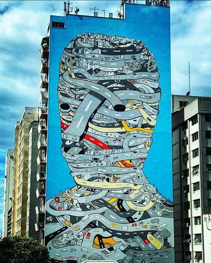 Street art wall. Huge mural on a 12-story skyscraper. Depicted is, on a blue background, a head with shoulders, which consists of endless streets. The streets snake across each other like gauze bandages and are populated with cars.