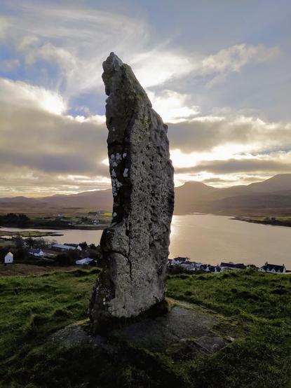 A large standing stone in the foreground overlooking a village and bay, with the sun illuminating a cloudy sky