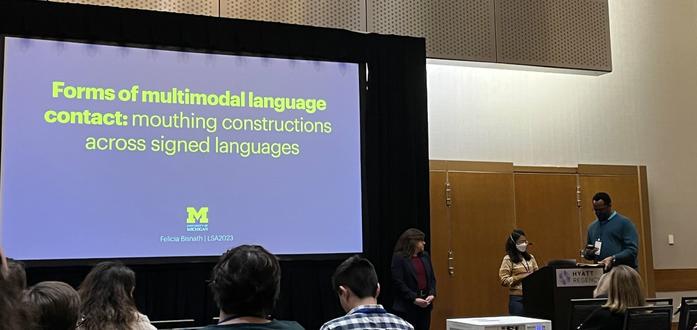 Presentation at the 2023 Linguistic Society of America conference. Picture shows view from audience. Text on screen: "Forms of multimodal contact: mouthing constructions". Presenter is a woman of Indian descent wearing a mask.