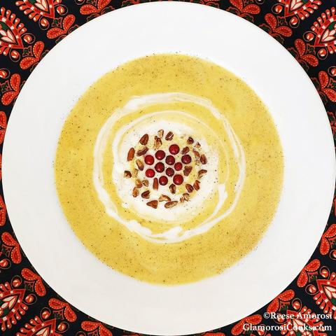 This square photo shows the completed Roasted Butternut Squash and Apple Soup recipe by Reese Amorosi for the Glamorosi Cooks website. The round bowl is white and the soup is pale orange. The soup is garnished with a swirl of maple creme fraiche, then topped with pecans, red currants and a dusting of nutmeg. The bowl is in closeup, but around the edges there is a a tablecloth with a black, red, orange and white printed design.