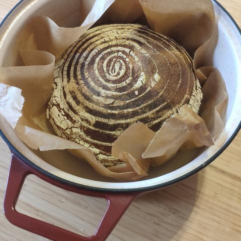 Ausgebackener runder Laib Weizenbrot mit brauner Kruste und typischer Gärkorbbemehlung auf Backpapier im gusseisernen Topf liegend.