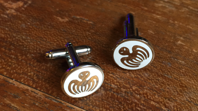 A pair of metal cufflinks lying on a wooden surface. They display the logo – a stylized #octopus – of the fictional organisation #SPECTRE from the #JamesBond franchise as a silver metal inlay on white background.