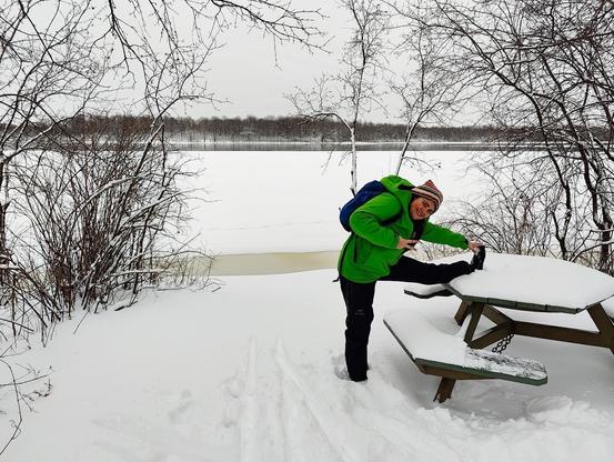 Devant le bord du fleuve, un paysage complètement enneigé et calme. De l'autre bord de la rive, une forêt dort sous la neige. Gersande porte une combinaison de ski vert et noir, s'étire la jambe sur une table do pique-nique, et fait le signe de la paix avec sa main droite.
