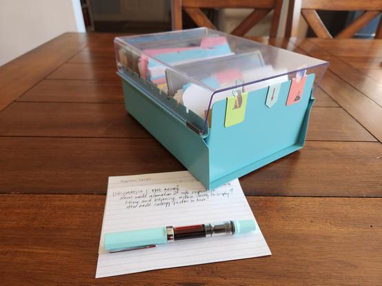 A small metal zettelkasten with a clear plastic lid sitting on a wooden table with a fountain pen and index card in front of it