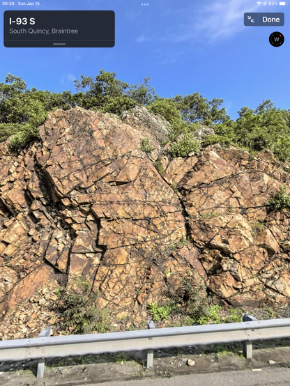 An exposure of the Mattapan along 1-93 S near the Quincy-Braintree boundary, showing characteristic orange hue and angular fractures. The freeway guardrail is visible at the bottom, and low trees appear below a blue sky. From Apple Maps.