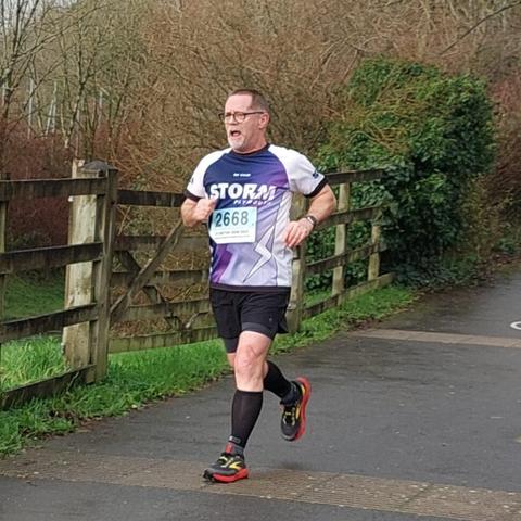 With about 15 metres to go and nothing left to give, I am the only person in the photo running on a paved surface beside a wooden fence. I am wearing red and grey trainers, black socks, black shorts and a purple and white short-sleeved running shirt with my number attached to the front under the word Storm which is the name of the running club which I belong to.