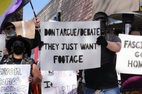 "Don't argue/debate. They just want footage" on poster board sign held up by person in the middle of the group of people who showed up to protect the event. Others are also holding signs, standing together in front of a building, wearing various sunglasses and face coverings.